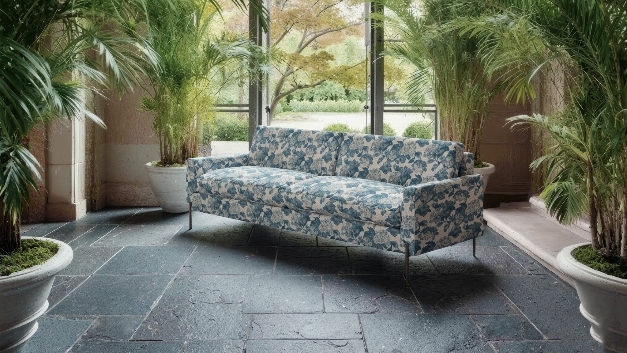 Decorative sofa with floral pattern in a room with large windows and potted plants.